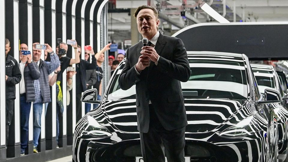 Elon Musk mit Mikro vor einem schwarzen Tesla bei einer Veranstaltung