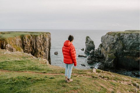Eine Frau steht an einer Steilküste und trägt eine rote Steppjacke.