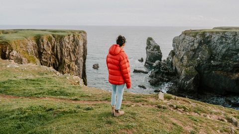 Eine Frau steht an einer Steilküste und trägt eine rote Steppjacke.