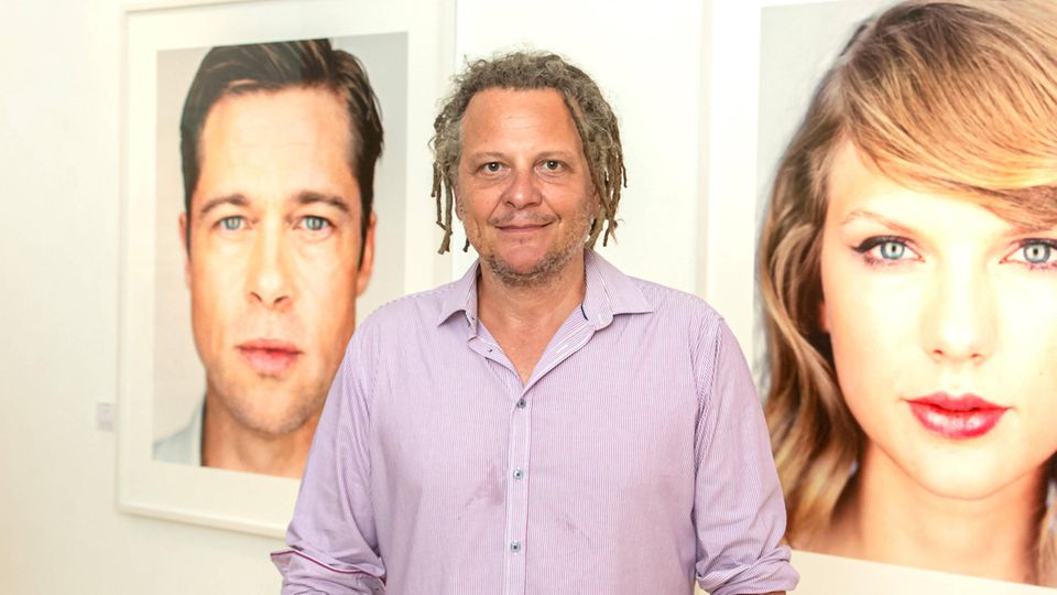 Martin Schoeller, ein Mann mit kurzen Dreadlocks, steht zwischen zwei seiner Fotos. Sie zeigen Brad Pitt und Taylor Swift