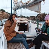 Ein Paar sitzt an Bord seines Segelschiffes, er trinkt Kaffee, sie arbeitet auf dem Laptop