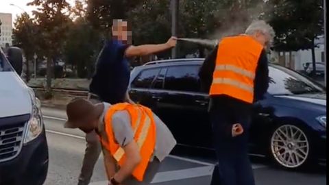 Ein Autofahrer steht auf der Straße und sprüht einem Klimaaktivisten in orangener Weste Reizgas ins Gesicht