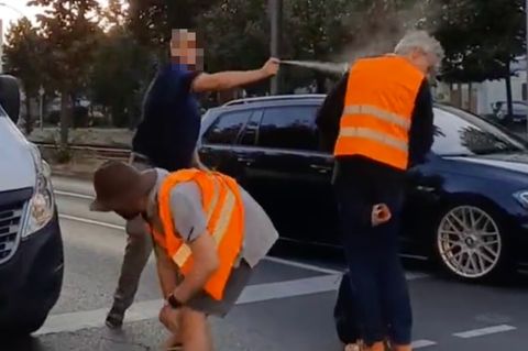 Ein Autofahrer steht auf der Straße und sprüht einem Klimaaktivisten in orangener Weste Reizgas ins Gesicht