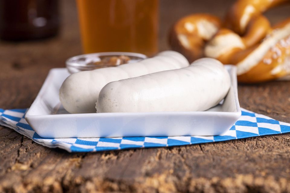 Zwei Weißwürste auf einem Teller, im Hintergrund liegt eine Brezel Zwei Weißwürste auf einem Teller, im Hintergrund liegt eine Brezel