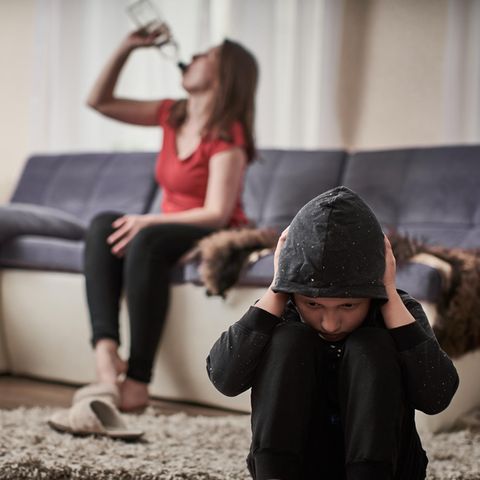 Eine Mutter sitzt auf dem Sofa und trinkt Alkohol, ein Kund sitzt davor und hält sich die Ohren zu
