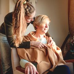 Eine junge Frau legt einer älteren Dame die an einem Fester sitzt eine Decke um die Schultern