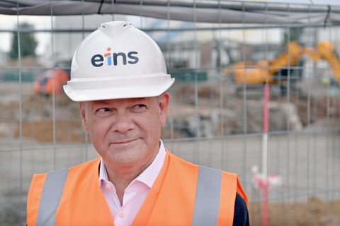 Olaf Scholz trägt auf einer Baustelle einen weißen Bauarbeiterhelm und eine orangene Warnweste