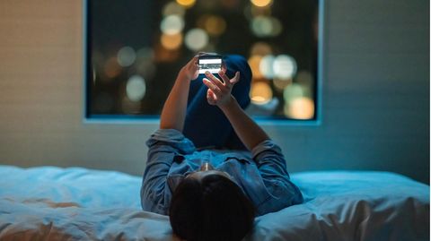 Eine Jugendliche liegt auf dem Rücken im Bett und sieht Fotos auf ihrem Handy an
