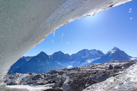 Von der Eiskante eines Gletschers tropft Schmelzwasser vor einer sonnigen Berglandschaft der Schweiz