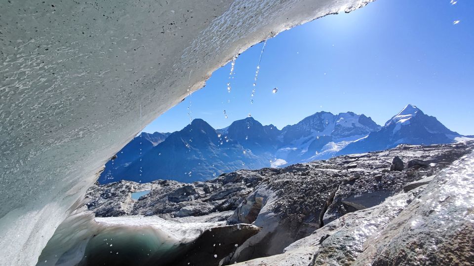 Von der Eiskante eines Gletschers tropft Schmelzwasser vor einer sonnigen Berglandschaft der Schweiz