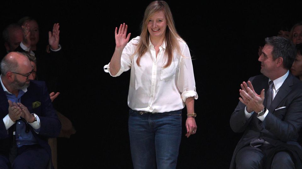 Sarah Burton, eine Frau in einer Bluse und in Jeans, winkt