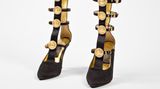 "Ein Aufeinandertreffen von Dschungel und Hofstaat", so beschrieb einmal die "New York Times" die Designs von Gianni Versace: leuchtende Farben einerseits und tiefes Schwarz andererseits, Riemen, Schnallen, metallische Akzente, der goldene Medusenkopf in seinem Logo sind Teil seiner Markenästhetik.   Gold, Riemen, Medusaköpfe – das Modell aus dem Jahr 1992 vereint alle Elemente, die für den Versace-Still so kennzeichnend sind.   Gianni Versace designte bis zu seinem gewaltsamen Tod im Jahr 1997. Dann übernahm seine Schwester Donatella die Leitung des Labels. Sie hatte bereits häufig mit ihrem Bruder zusammengearbeitet und präsentierte nur zwölf Wochen nach dessen Tod ihre erste Kollektion. Für ihren eigenen Stil griff sie Aspekte seiner Designphilosophie auf und entwickelte die Versace-Ästhetik weiter. "Manche meinten, Giannis Mode sei vulgär ... aber Frauen lieben sie nun mal, nicht weil sie Haut zeigte, sondern weil er die Frauen stark machte. Er sagte ihnen, sie sollten keine Angst vor ihrer Sexualität haben", erklärte Donatella. 