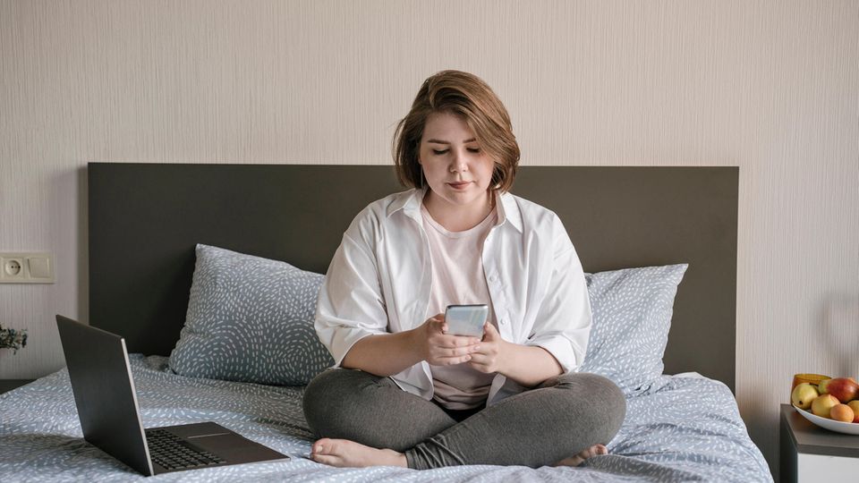 Junge Frau sitzt auf Bett und schaut auf Handy. Auf Tiktok wird viel über psychische Krankheiten gesprochen