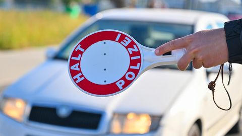 Eine linke Hand stoppt mit einer roten Kelle mit "Halt Polizei"-Schrift einen weißen Kleinwagen