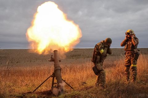 Eine Waffe feuert ab und zwei Soldaten stehen daneben und halten sich die Ohren zu