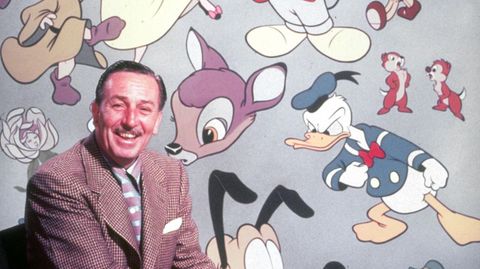 Walt Disney sitzt vor einer Wand mit den berühmtesten Disney-Figuren