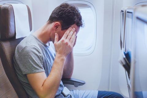 Fliegen: Ein Reisender sitzt in einem engen Flugzeug und hält sich die Hände vors Gesicht