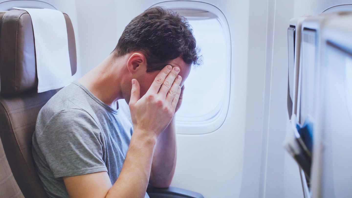 Fliegen: Ein Reisender sitzt in einem engen Flugzeug und hält sich die Hände vors Gesicht