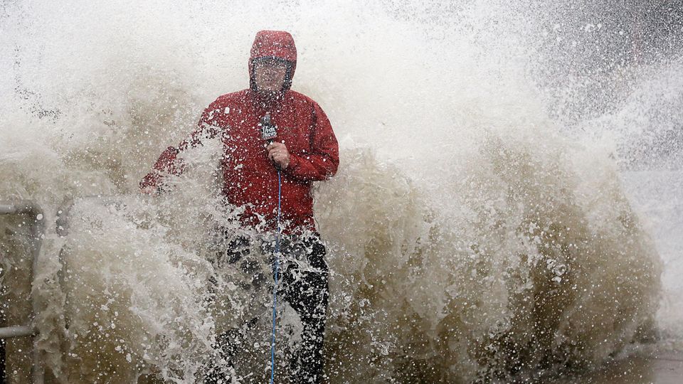 Ein Reporter in roter Jacke wird von einer seitlichen Welle überrascht