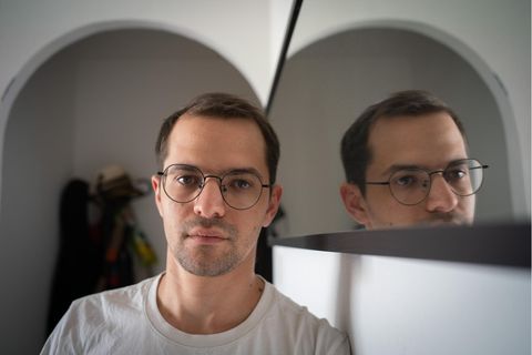 Benzion Sanders, braune kurze Haare mit Brille und weißem T-shirt, neben einem Spiegel