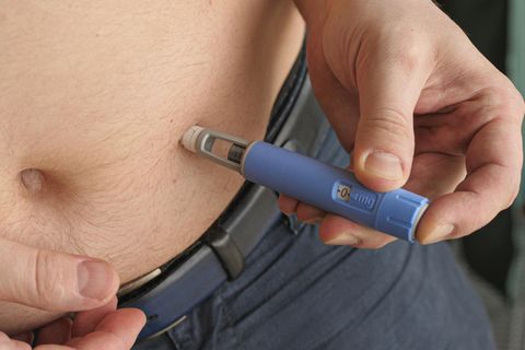 Ein Mann mit dunkler Hose setzt sich mit der Hand den hellblauen Ozempic-Injektionspen auf den nackten Bauch