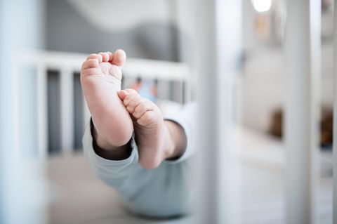 Durch die weißen Gitterstäbe eines Kinderbettchens sind die Füße eines Babys zu sehen, die aus einem hellblauen Strampler ragen