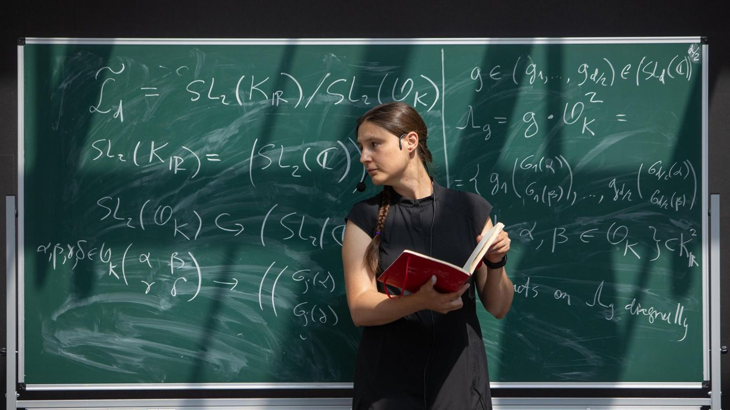 Maryna Viazovska steht mit einem Buch an einer Tafel voller Formeln