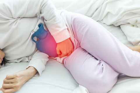 Eine Frau liegt leicht gekrümmt im weißen Schlafanzug im Bett und hält sich eine Wärmflasche vor den Unterleib
