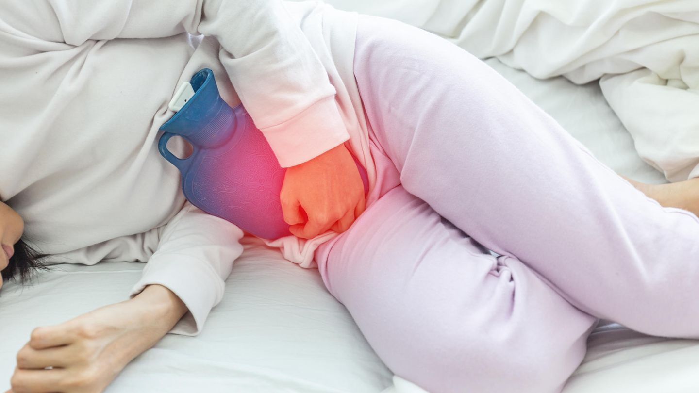 Eine Frau liegt leicht gekrümmt im weißen Schlafanzug im Bett und hält sich eine Wärmflasche vor den Unterleib Eine Frau liegt leicht gekrümmt im weißen Schlafanzug im Bett und hält sich eine Wärmflasche vor den Unterleib