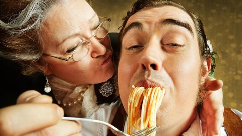 Eine Italienische Mama küsst ihren Erwachsenen Sohn auf die Wange während sie ihn mit Spagetti füttert
