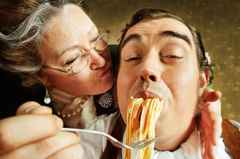 Eine Italienische Mama küsst ihren Erwachsenen Sohn auf die Wange während sie ihn mit Spagetti füttert