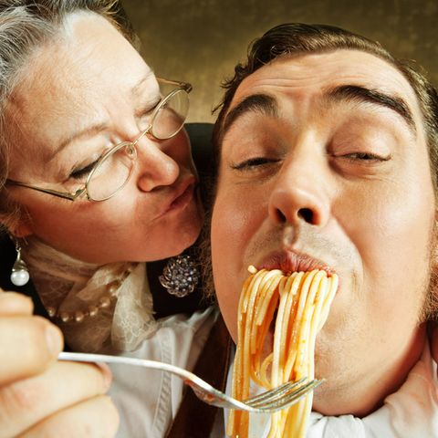 Eine Italienische Mama küsst ihren Erwachsenen Sohn auf die Wange während sie ihn mit Spagetti füttert