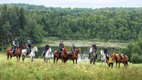 Eine Gruppe von Reitern mit Pferden steht vor einem See, dahinter ein Wald
