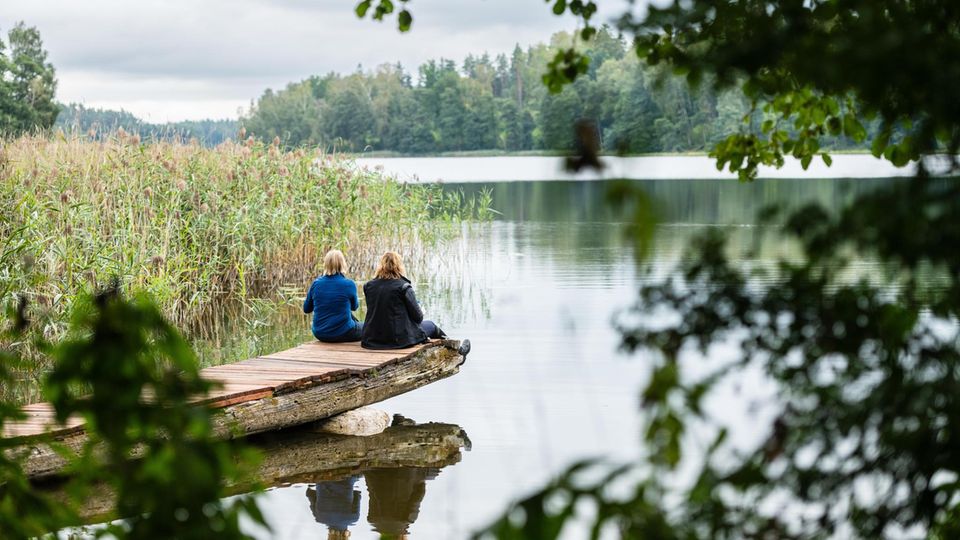 zwei Frauen mit dem Rücken zur Kamera sitzen auf einem Steg an einem See zwei Frauen mit dem Rücken zur Kamera sitzen auf einem Steg an einem See