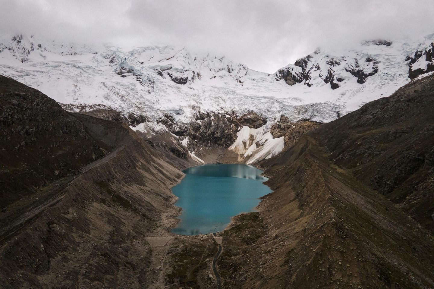 Ein blauer Gletschersee neben braunen Flächen, dahinter Eis