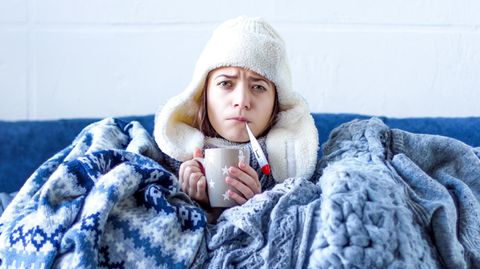 Frau mit Erkältung sitzt mit Wolldecke und Mütze auf dem Sofa