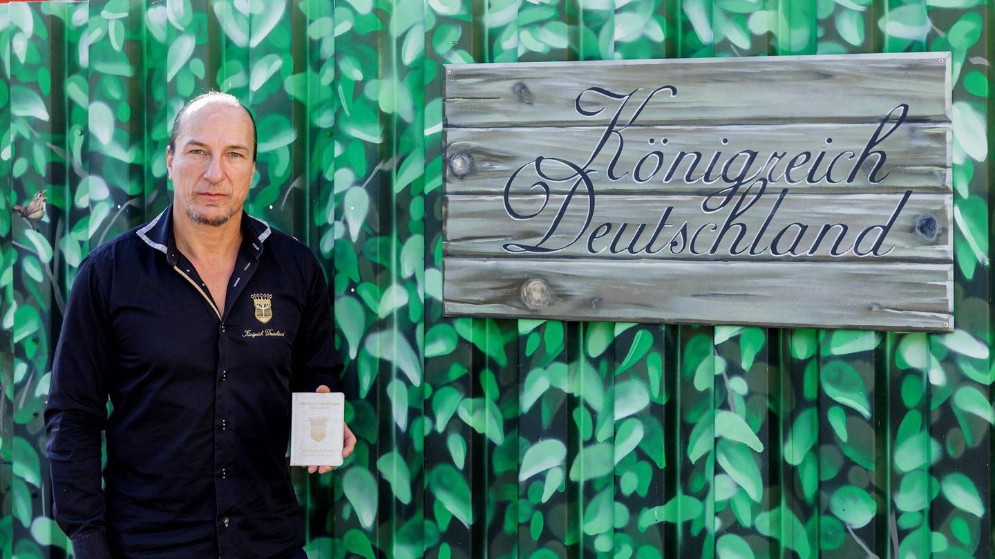 Peter Fitzek steht mit einem Ausweis in der linken Hand neben einem Schild "Königreich Deutschland"