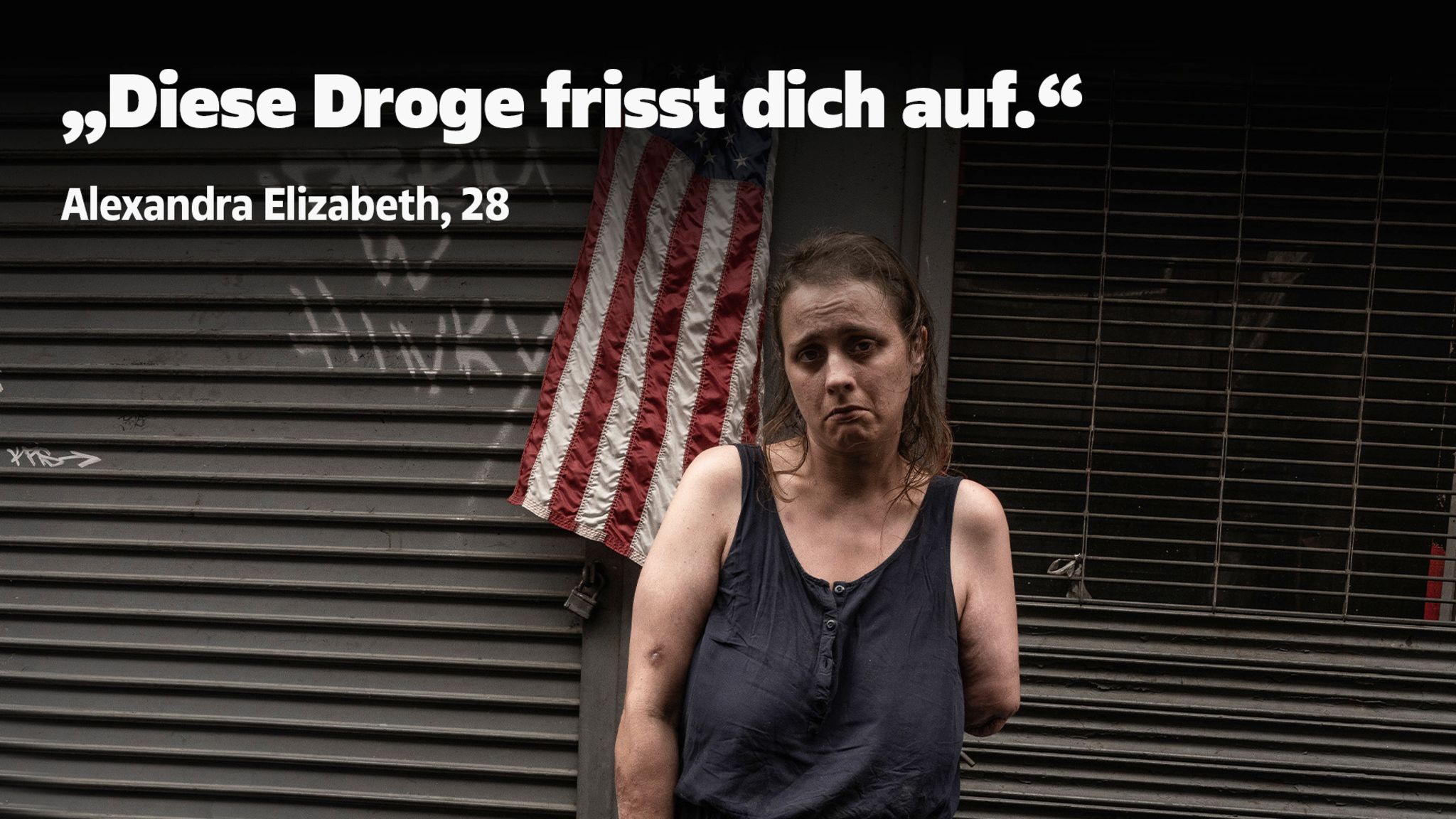Eine Frau mit einem amputierten Arm steht vor einer Wand an der eine USA-Flagge hängt und guckt traurig