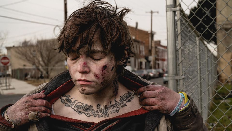 Eine junge Frau mit starken wunden im Gesicht zieht ihre Kleidung zur Seite um ein Tattoo zu zeigen ihre Augen sind geschlossen