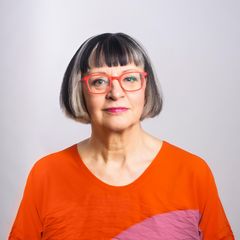 Eine Frau mit kinnlangem dunklem Haar mit Pony und orangefarbener Brille lächelt in die Kamera