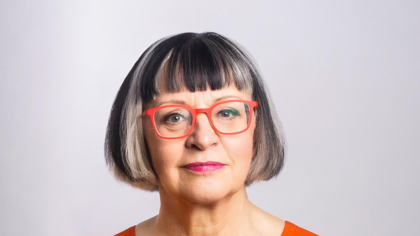 Eine Frau mit kinnlangem dunklem Haar mit Pony und orangefarbener Brille lächelt in die Kamera