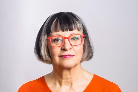 Eine Frau mit kinnlangem dunklem Haar mit Pony und orangefarbener Brille lächelt in die Kamera