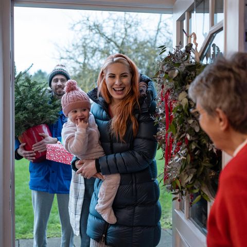 Eine Familie mit Kind kommt bei der Oma zu Weihnachten durch die Tür und bringt einen Tannenbaum mit