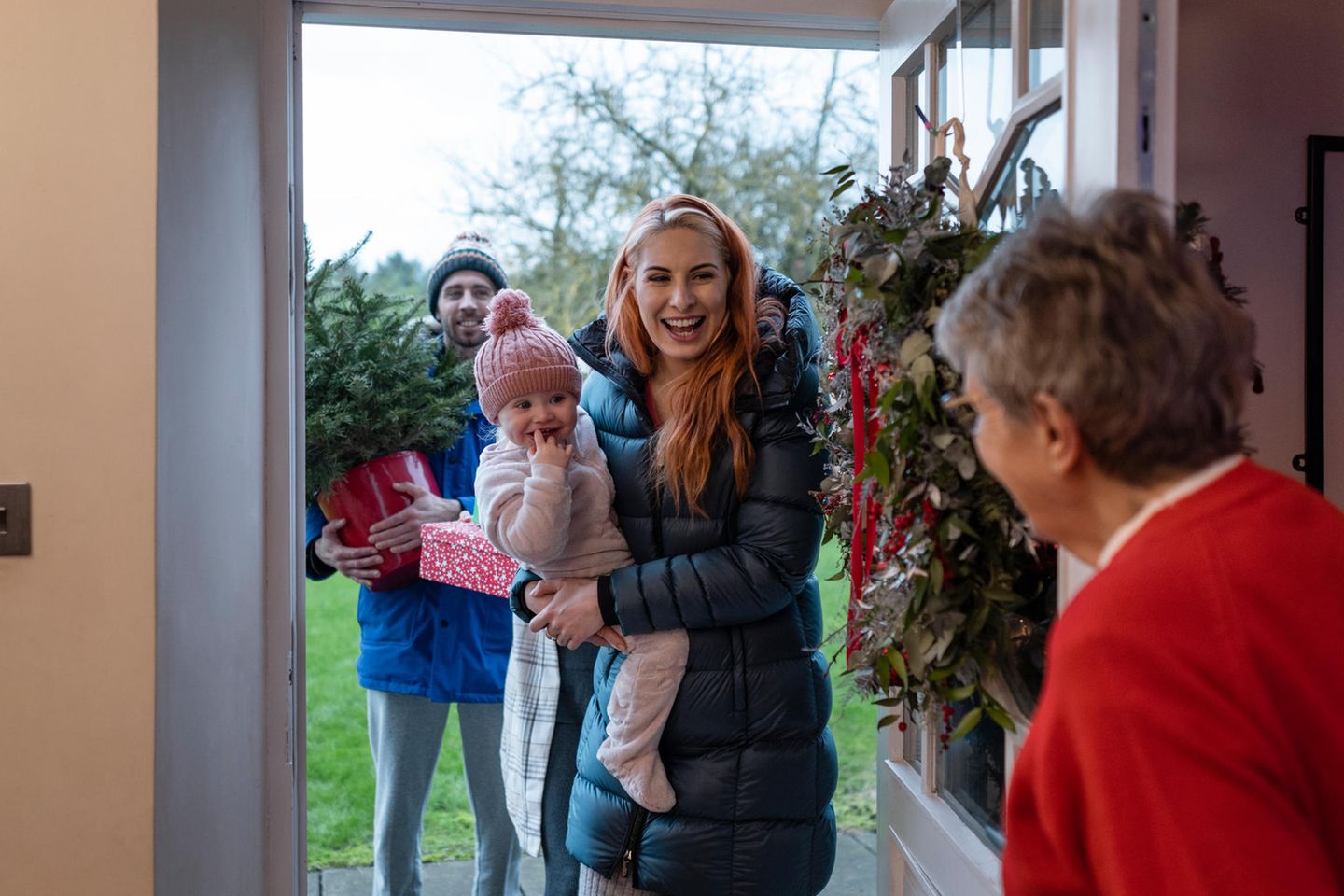 Eine Familie mit Kind kommt bei der Oma zu Weihnachten durch die Tür und bringt einen Tannenbaum mit
