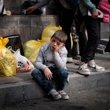 Ein verletzter junge sitzt auf einem Bordstein zwischen gepackten Plastiktüten und stützt seinen Kopf auf seine Hände