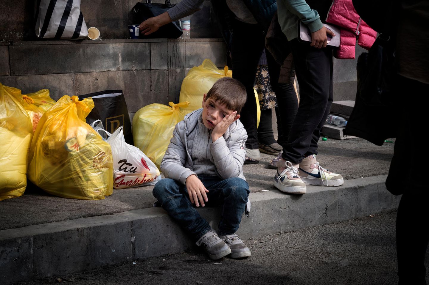Ein verletzter junge sitzt auf einem Bordstein zwischen gepackten Plastiktüten und stützt seinen Kopf auf seine Hände