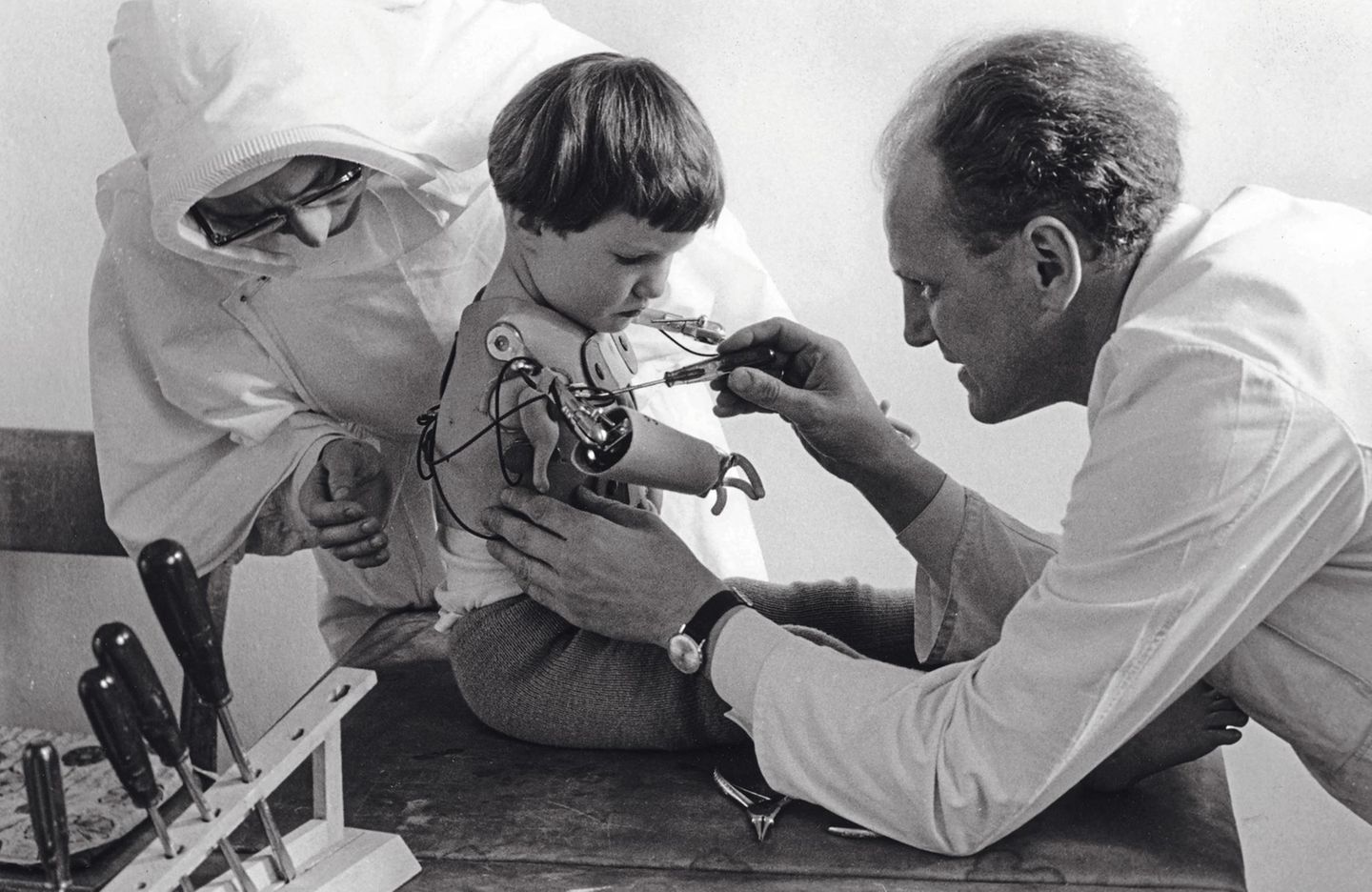 1965 - Neuwied Nebenwirkungen Ist dies ein guter, hoffnungsvoller Tag? Oder wieder ein Tag, an dem das Kind schmerzlich spürt, dass es anders ist als die anderen? Ein Arzt und eine Schwester passen dem Mädchen in einem spezialisierten Kinderheim in Neuwied eine Arm-prothese an. Fehlbildungen an Armen und Beinen gehören zu den schweren Schädigungen, die der Wirkstoff Thalidomid hervorgerufen hatte – enthalten im Schlaf- und Beruhigungsmittel Contergan. Das anfangs als „gut verträglich“ bezeichnete Medikament, das seit 1957 rezeptfrei erhältlich ist, war nicht an Schwangeren getestet worden. Aber schon eine einzige Tablette genügt, um dem heranwachsenden Kind im Mutterleib schwer zu schaden. Allein in Deutschland kommen so etwa 5000 Kinder mit Missbildungen zur Welt. 40 Prozent von ihnen sterben kurz nach der Geburt oder im Säuglingsalter. Ein Prozess gegen die Herstellerfirma Grünenthal wird 1970 eingestellt. Die Opfer des Skandals erhalten Entschädigungszahlungen aus einem Stiftungsfonds, der von Grünenthal und dem Bund bestückt wird. 1997 ist das Geld aufgebraucht. Seitdem zahlt der Bund die Contergan-Renten weitgehend allein.