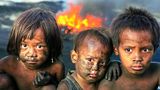 Drei mit Ruß verschmierte Kinder asiatischer Abstammung schauen in die Kamera, hinter ihnen eine Flamme