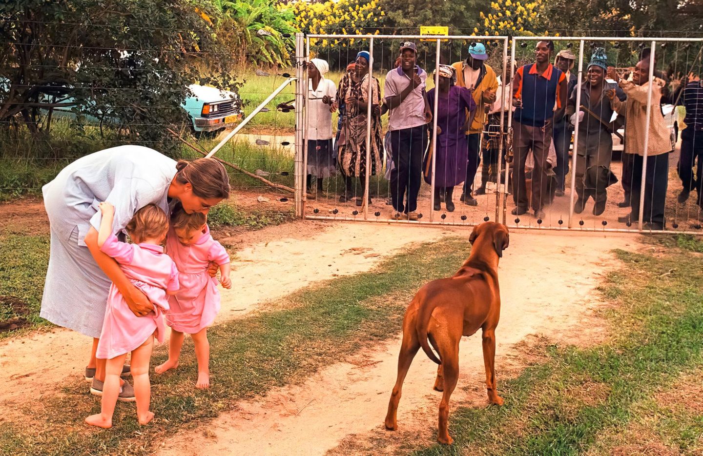 2000 – Simbabwe Simbabwe: Frau mit zwei Töchtern und Hund sieht sich erbosten Menschen hinter einem Zaun gegenüber