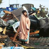 Eine junge Frau steht in eine Decke gewickelt inmitten von Trümmern.